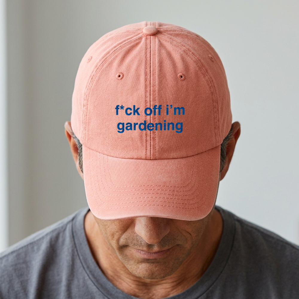 A male model stood in front of a blurred background wearing a premium vintage embroidered cap in vintage peach reading f*ck off i'm gardening in blue custom text 