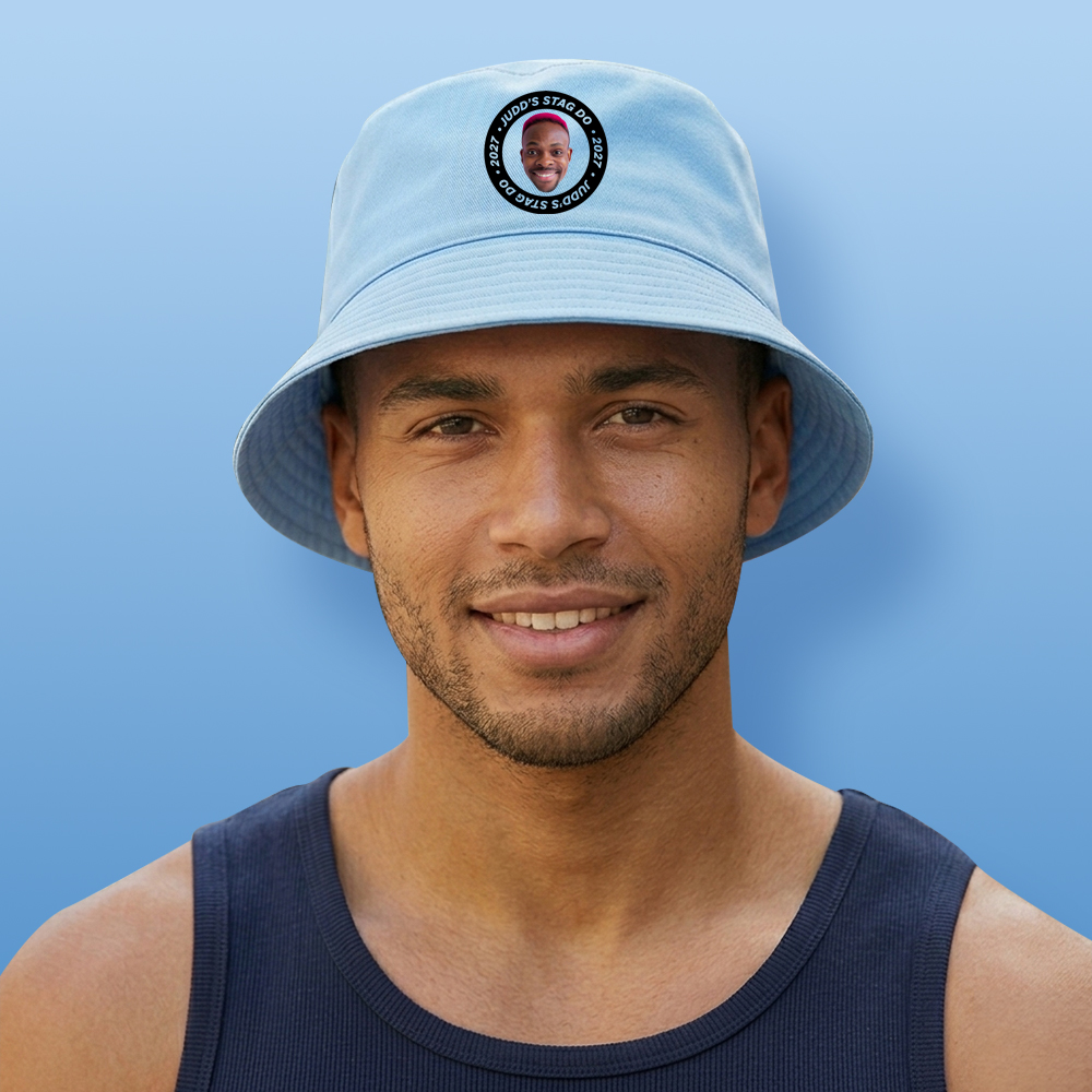 A male model wearing a light blue stag do bucket hat that has a circular football badge style design that has been personalised with a photo of a man and the text Judd's Stag Do 2027