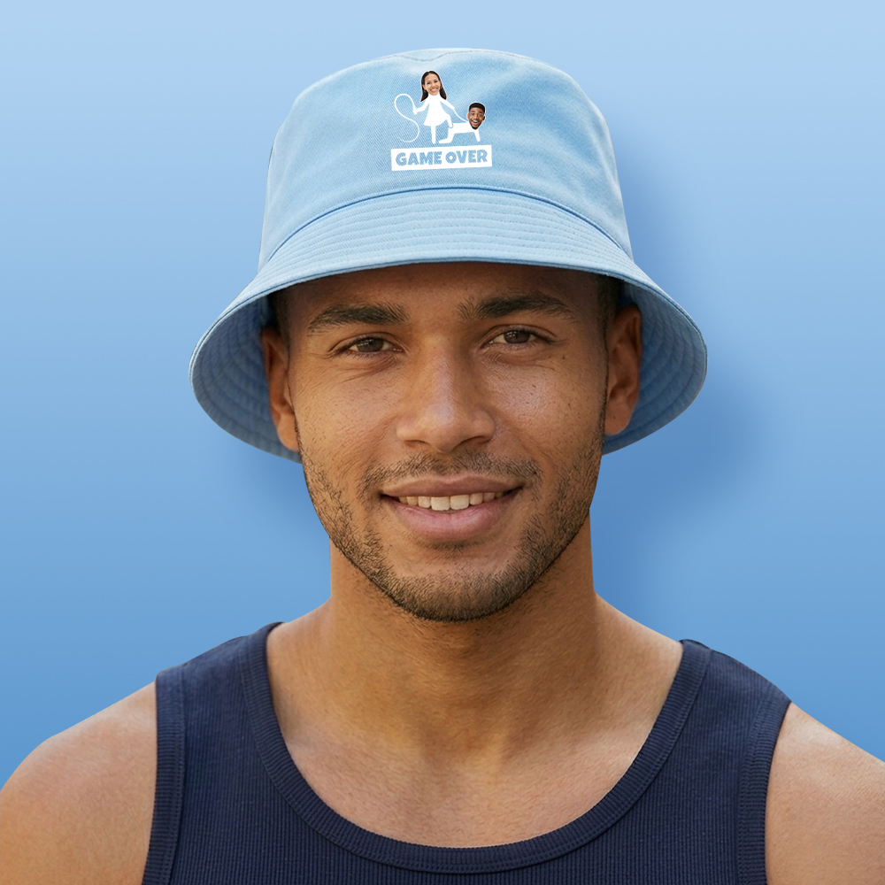 A male model wearing a light blue custom stag do bucket hat where you can upload two face pics to make it look like the stag is being whipped by his partner with the text Game Over