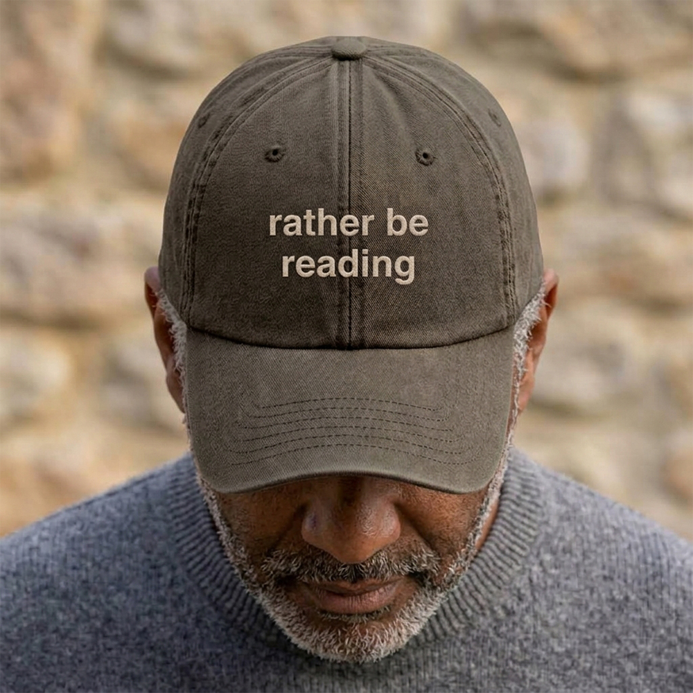 A male model stood in front of a blurred background wearing a premium vintage embroidered Father's day cap in brown with custom text reading rather be reading in tusk font