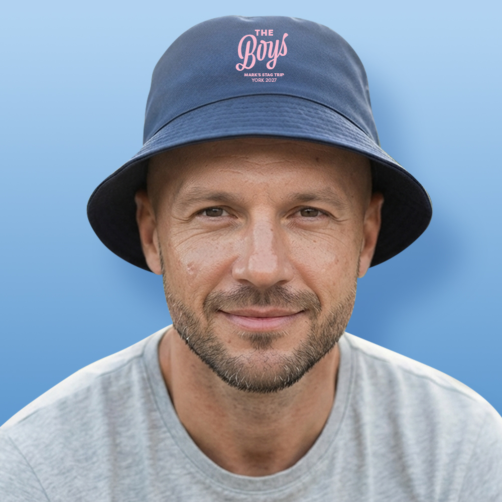 A male model wearing a light blue do custom bucket hat that reads The Boys in a fancy handwriting font with custom text Mark's Stag Trip York 2027 underneath it