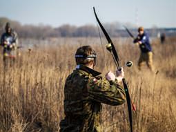 Image of men in a field with archery bows aiming to fire arrows at each other