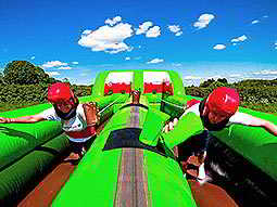 Two people running on a green bouncy castle with red helmets on, in a field 