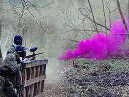Two people hiding behind a fence and playing paintball with pink smoke in the foreground
