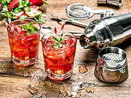 Two red cocktails topped with ice, leaves and straws, next to a cocktail shaker