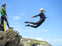 A man jumping off a cliff in a wetsuit, whilst someone looks on at Coasteering in Newquay