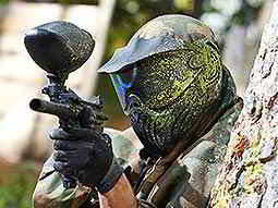 A man wearing a paintball mask covered in yellow paint aims a paintball gun from behind a tree