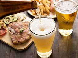 A glass of beer being filled from a bottle, with another glass of beer, a steak meal and a plate of chips