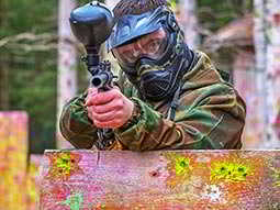A man wearing a paintball mask standing behind a wooden board and aiming a paintball gun