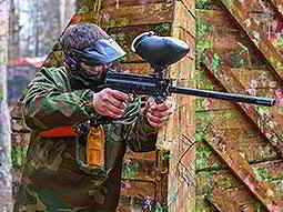 A man standing next to a wooden barricade, holding his paintball gun