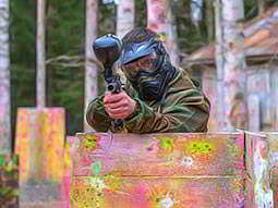 A man aiming his paintball gun at the camera, whilst sheltering his lower half with a wooden barrier