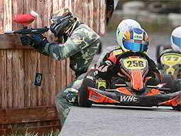 A split image of a man aiming his gun through a hole in a fence and some people driving go karts around a track