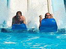 Two men landing in a swimming pool on blue floats, at the end of a slide