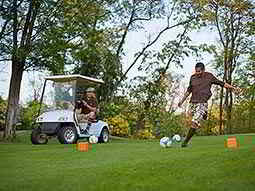 A man in golf attire, kicking a football on a golf course with a man sat in a golf buggy in the background