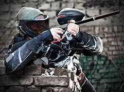 A man wearing a paintball mask leaning on a brick wall and aiming a paintball gun