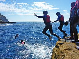 Two people jumping into water off a rock, with a group of people watching