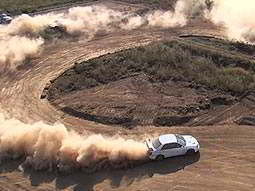 A white car driving around a hairpin bend on a dirt road, kicking up dust behind it