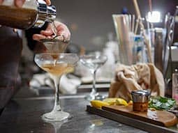 A man's hand pouring a cocktail through a shaker and into a martini glass, with utensils and ingredients on the side