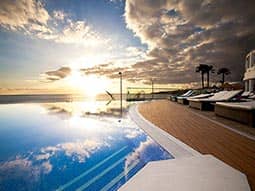 A pool surrounded by sun loungers, to a backdrop of the coast