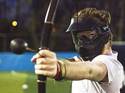 A split image of a man with a paintball splatter on his visor, and a man firing a bow and arrow at a target