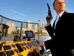 A man in a black suit, holding a handgun whilst driving a boat 