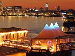A marquee connected to a lit-up boat on the River Rhein, with buildings in the background