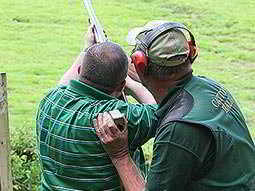 A man aiming with a shotgun to the sky, as an instructor looks on and holds his arm