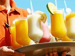 A male bartender in an orange polo, holding up a tray of colourful cocktails