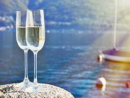 Close up of two flute glasses filled with champagne on a rock with a small yacht and boys on water in the background