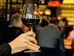 A close up focusing of someone holding a glass of red wine, with some people in the background