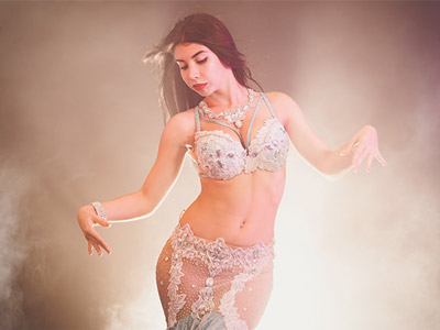 A woman belly dancing with a silver outfit on and smoke behind her