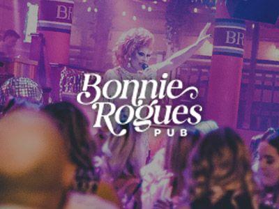 A drag queen with a red wig talking into a microphone wearing a white dress with the Bonnie Rogues Pub logo layered over it 