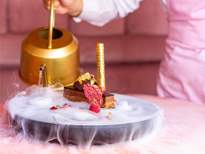 A person pouring some tea from a gold kettle onto a platter that has a piece of millionaire's shortbread on it 