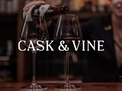 Someone pouring some red wine into two wine glasses with the Cask & Vine logo layered over it 