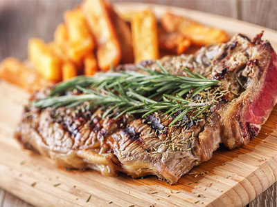 A steak on a wooden board with chips on a plate in the background and a drink being poured