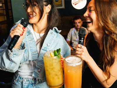 Two women singing karaoke and drinking large colourful cocktails 