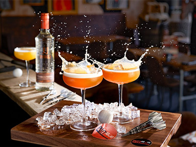 A wooden board with two orange cocktails, a mini golf ball and some darts 