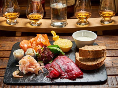 A black slab with some food including salmon, prawns and bread on it sat next to a tasting tray of whiskies 