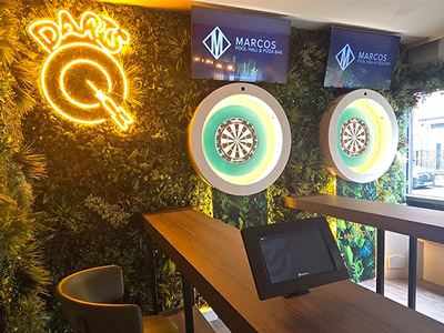 Two darts oches next to each other at Marcos Pool Hall with a yellow neon sign of a darts board reading Darts 