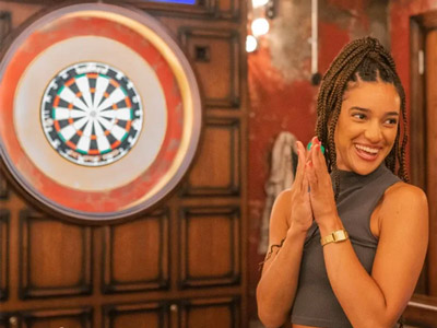 A smiling woman with her hands clapped together stood in front of a darts board 