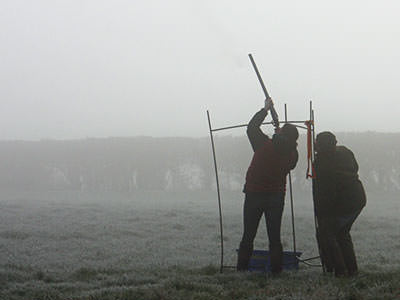 A close up of a man firing a shotgun to the sky, with someone else looking on to a fooggy backdrop