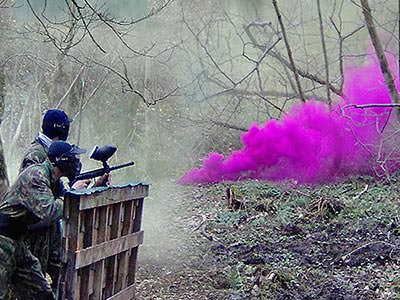 Two people hiding behind a fence and playing paintball outdoors with pink smoke in the foreground