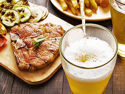 A steak on a wooden board with chips on a plate in the background and a drink being poured