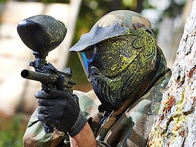 A man wearing a paintball mask covered in yellow paint aims a paintball gun from behind a tree