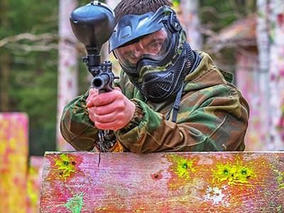 A man wearing a paintball mask standing behind a wooden board and aiming a paintball gun