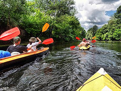 The back of two canoes full of people, sailing down a river with trees on either side
