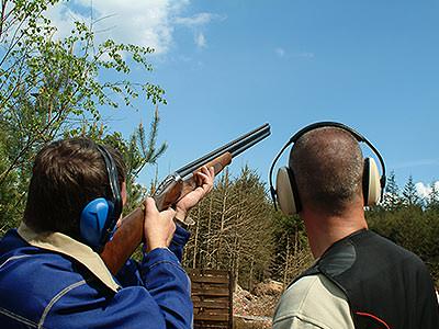 The back of a man aiming to the sky with a shotgun, with an instructor stood to the side of him