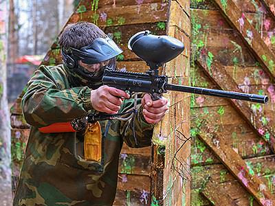 A man standing next to a wooden barricade, holding his paintball gun