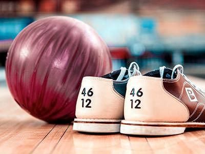 A pair of bowling shoes next to a red bowling ball, with the bowling alley in the background