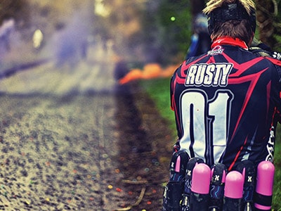 A man firing a paintball gun with 'Rusty' on his shirt